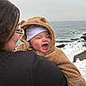 baby, child, woman, glasses, ocean, snow, winter_clothing, hoodie, smile, outdoor, coastline, cold_weather, happy, person, face, water, nature, rock, sky, holding