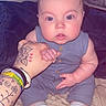 baby, infant, child, hand, tattoo, blue_romper, striped_socks, bed, blanket, adult_hand, fan, indoor, cute, wide_eyes, skin, person, holding, sitting, comfort, curious
