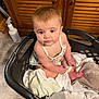 baby, child, laundry_basket, towels, clothes, floor, wooden_cabinet, indoor, person, sitting, curious, cute, infant, skin, hair, eyes, hands, feet, fabric, home