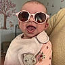 baby, sunglasses, tongue_out, bib, floral_clothing, indoor, smiling, face, child, cute, person, decor, wall_art, tree_mural, butterflies, pacifier_clip, handheld, pink, soft_texture, happy