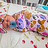 animal_print, baby, baby_supplies, bedding, changing_mat, cherry_pattern, child, colorful_clothing, crib, diapers, headband, indoor, infant, lying_down, nursery, pacifier, pink_outfit, portrait, swaddled, wide_eyed