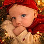 baby, child, blue_eyes, red_headband, bow, hand_in_mouth, holiday_lights, pine_branches, festive, cozy, warm_colors, sweater, heart_pattern, close_up, portrait, cute, infant, christmas_theme, soft_texture, snuggled