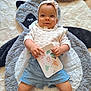 baby, child, headband, knitted_clothing, blue_shorts, plush_mat, animal_mat, card, indoor, floor, cute, smiling, person, infant, soft_texture, lying_down, handheld_object, decor, happy, portrait