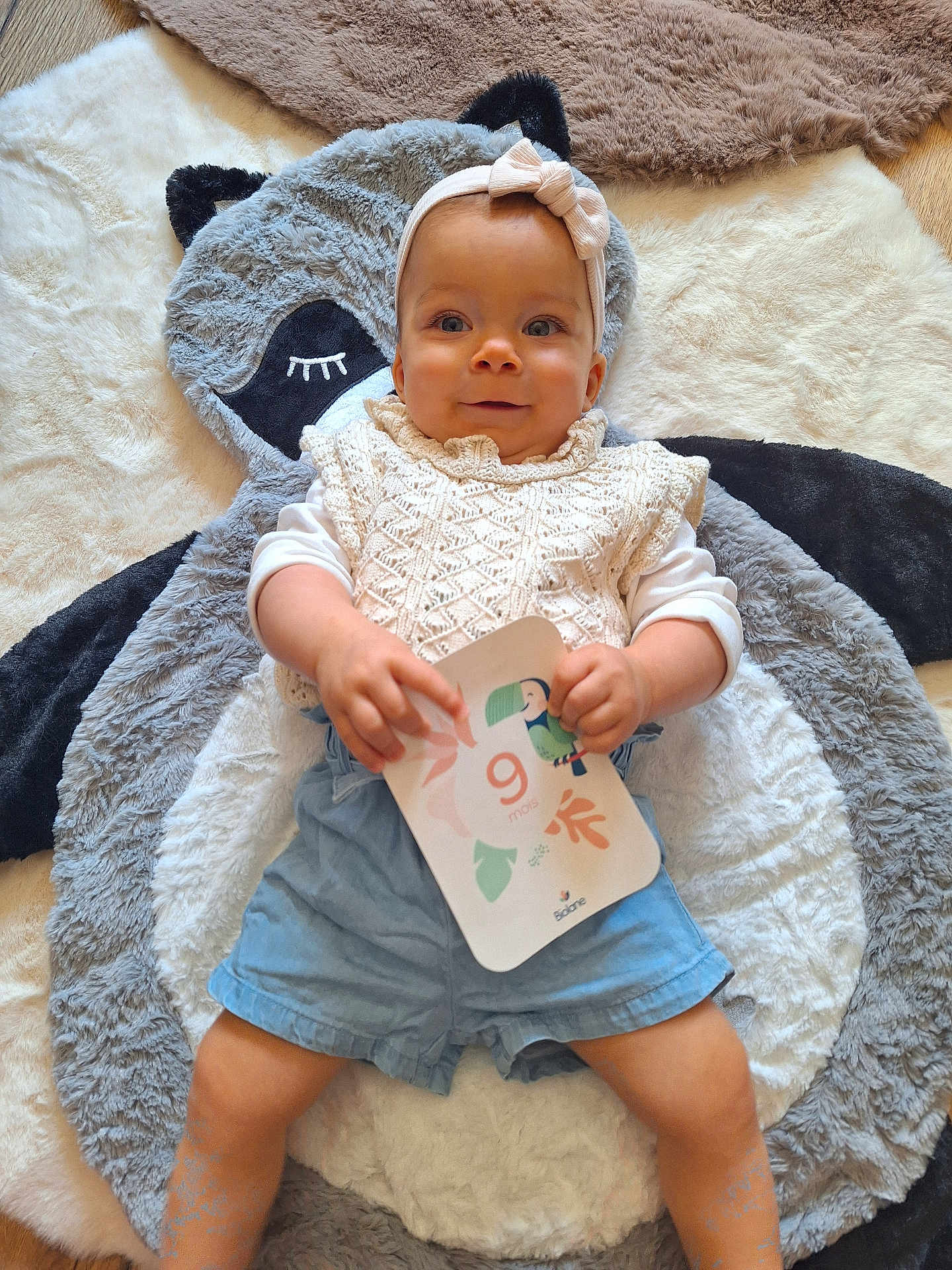Aria participe au concours pour gagner de l'argent avec cette photo : baby, child, headband, knitted_clothing, blue_shorts, plush_mat, animal_mat, card, indoor, floor, cute, smiling, person, infant, soft_texture, lying_down, handheld_object, decor, happy, portrait