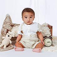Abiel joined the competition — help win amazing prizes! baby, child, sitting, white_background, wooden_bed, crocheted_toy, alarm_clock, beige_clothing, suspenders, barefoot, neutral_colors, soft_fabrics, portrait, indoor, cute, infant, baby_boy, toy, decor, floor