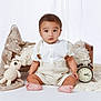 baby, child, sitting, white_background, wooden_bed, crocheted_toy, alarm_clock, beige_clothing, suspenders, barefoot, neutral_colors, soft_fabrics, portrait, indoor, cute, infant, baby_boy, toy, decor, floor