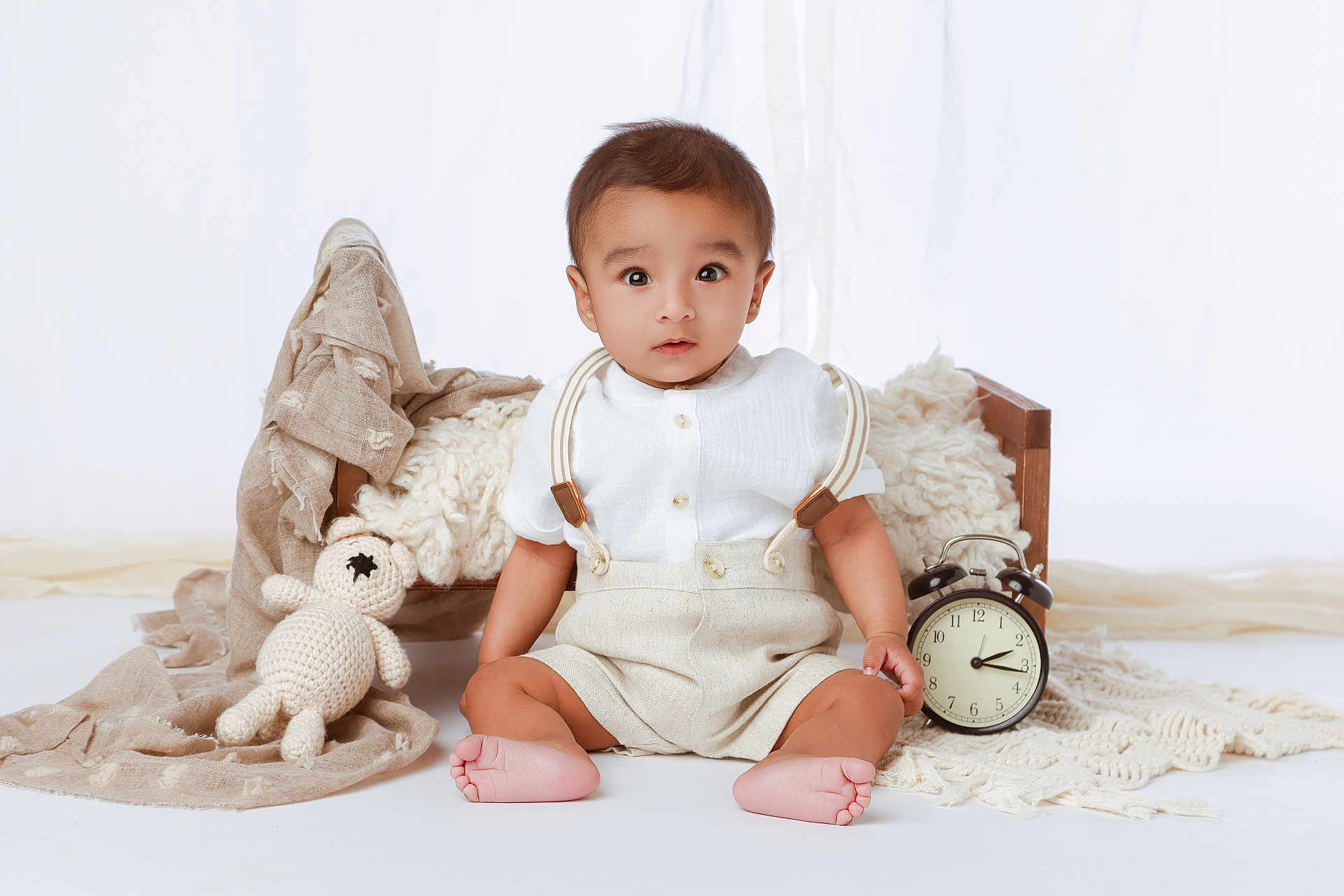 Abiel joined the competition — help win amazing prizes! baby, child, sitting, white_background, wooden_bed, crocheted_toy, alarm_clock, beige_clothing, suspenders, barefoot, neutral_colors, soft_fabrics, portrait, indoor, cute, infant, baby_boy, toy, decor, floor