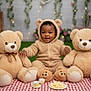 baby, child, teddy_bear, plush_toy, picnic_blanket, green_grass, cute, smiling, onesie, hooded_clothing, indoor, decorative_lights, flowers, playful, sitting, soft_toys, cozy, portrait, happy, infant