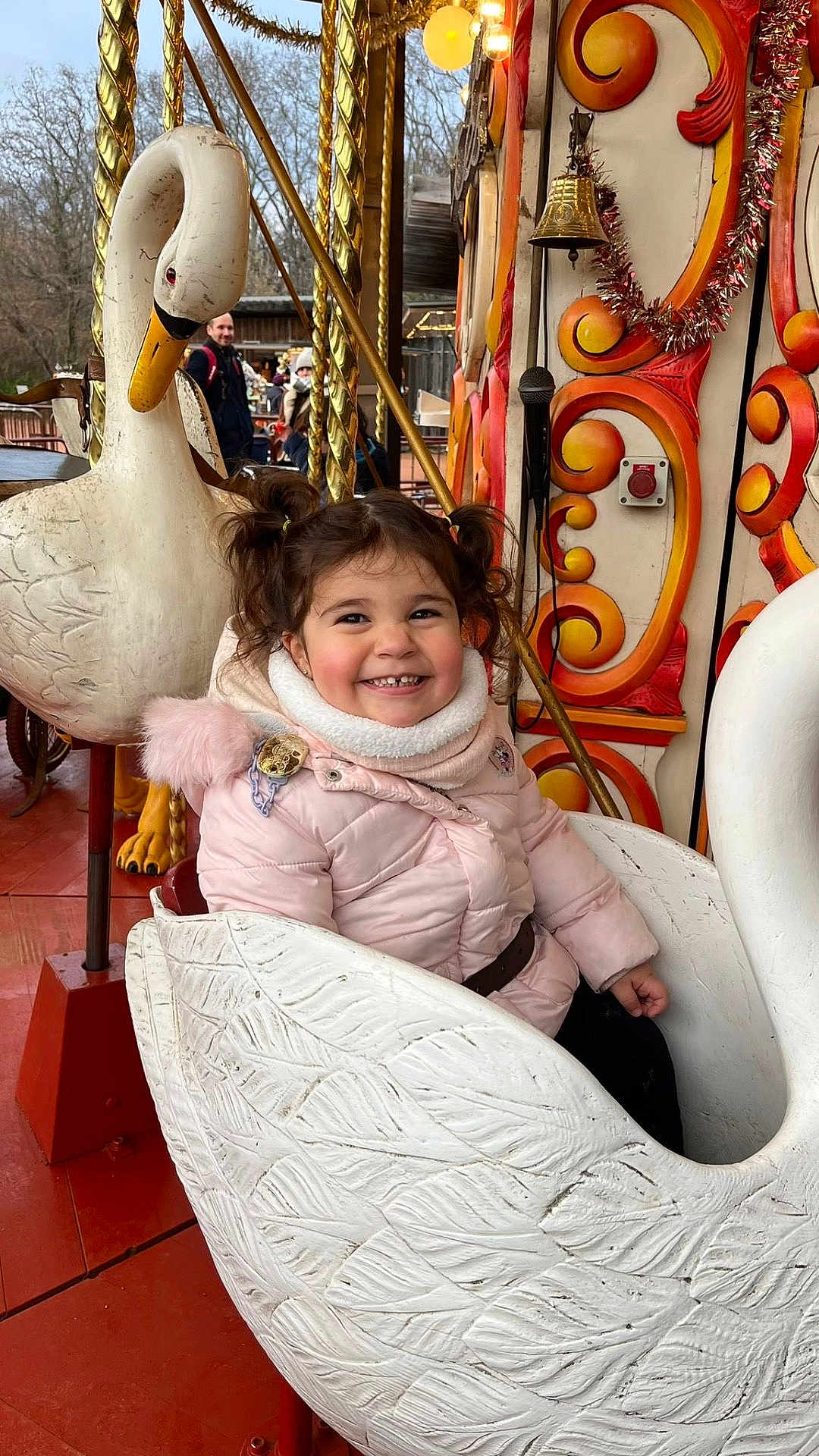 Luna participe au concours pour gagner de l'argent avec cette photo : child, toddler, girl, smile, carousel, swan, amusement_ride, pigtails, winter_coat, outdoor, seat, ornate_decoration, happy, playful, family, belt, wooden_floor, bell, person_in_background, portrait