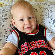 Kerem joined the competition — help win amazing prizes! baby, child, smiling, blue_eyes, jersey, black_clothing, red_text, white_shirt, cushion, patterned_cushion, indoor, portrait, baby_face, cute, infant, happy, reaching_out, light_skin, short_hair, closeup