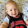 baby, child, smiling, blue_eyes, jersey, black_clothing, red_text, white_shirt, cushion, patterned_cushion, indoor, portrait, baby_face, cute, infant, happy, reaching_out, light_skin, short_hair, closeup