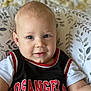baby, child, face, blue_eyes, smile, jersey, clothing, los_angeles, black, red, white, sleeves, cushion, pattern, leaves, indoor, portrait, cute, infant, seated