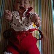 Ezio participe au concours pour gagner de l'argent avec cette photo : baby, child, smiling, reaching, red_shorts, white_shirt, patterned_clothing, striped_background, happy, playful, indoor, portrait, infant, cute, person, clothing, fabric, colorful, young_child, lying_down