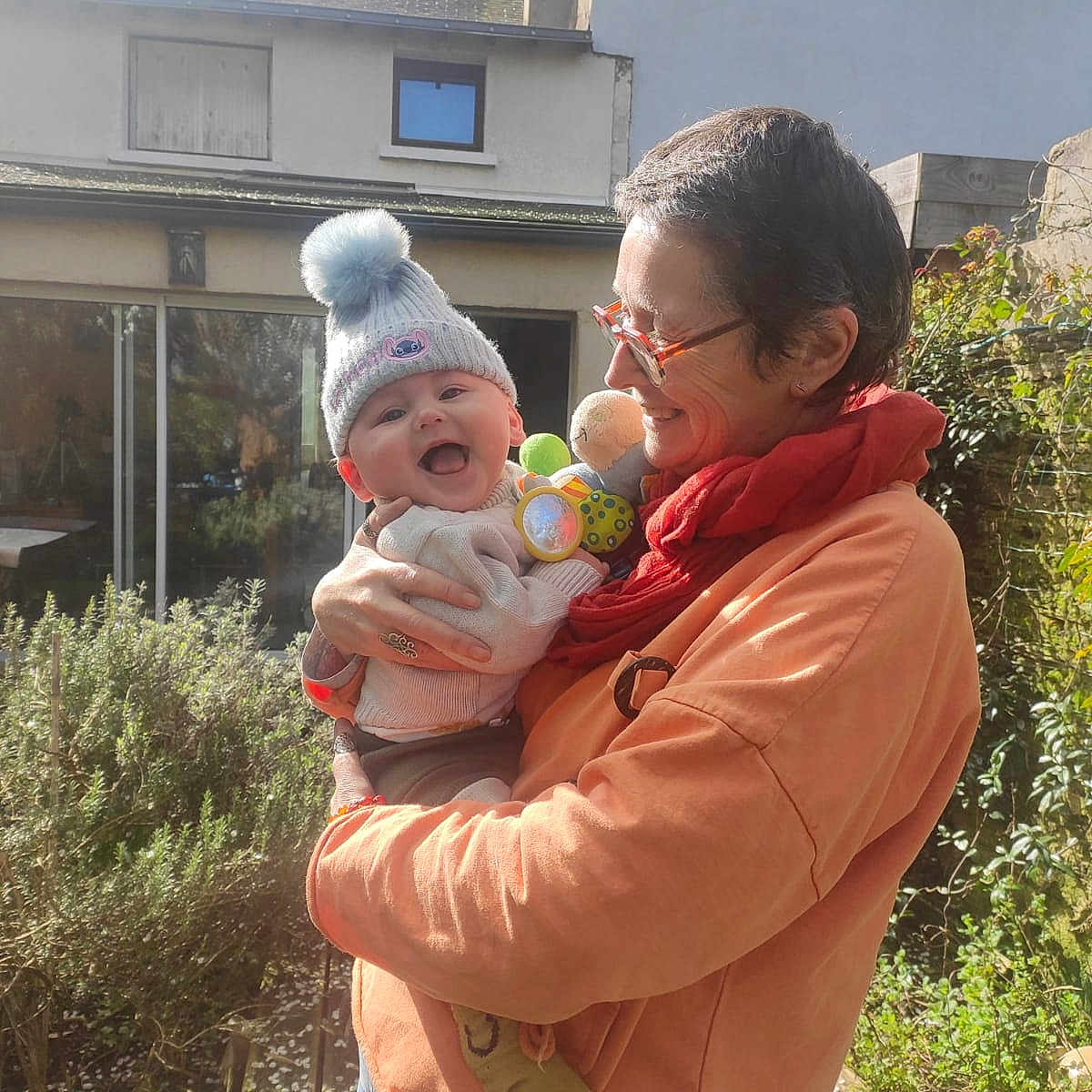 Ezio participe au concours pour gagner de l'argent avec cette photo : baby, child, adult, person, outdoor, garden, hat, pom_pom, scarf, smile, happy, holding, clothing, greenery, sunlight, house, window, plants, portrait, daylight
