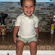 Kymir is registered to the contest to win money with this photo: child, toddler, smiling, happy, indoor, kitchen, counter, shorts, shirt, footwear, utensils, can, spray, tile_wall, cabinet, person, face, curly_hair, sitting, appliance