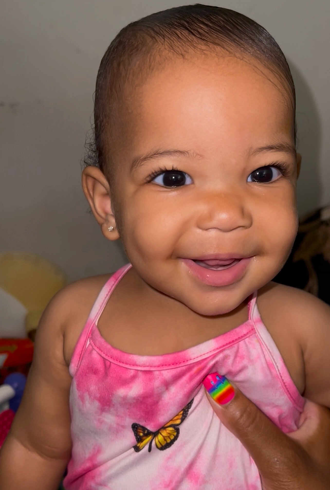 Avierra is registered to the contest to win money with this photo: baby, smiling, child, face, pink_clothing, earrings, hand, rainbow_nails, butterfly, tank_top, skin, portrait, cute, happy, indoor, closeup, person, infant, young_child, joyful