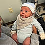 baby, infant, person, clothing, mitten, hat, holding, indoor, medical_equipment, power_outlet, face, expression, curious, seated, adult_hand, sleeve, blanket, floor, footwear, headwear