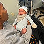 baby, infant, person, adult, clothing, hat, mittens, gray_sweatshirt, hospital, medical_equipment, chair, floor, power_outlet, blanket, seated, holding, indoor, curious, cute, warm