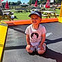 toddler, child, trampoline, cap, smile, sunlight, outdoor, netting, picnic_table, umbrella, grass, summer, playground, shorts, tshirt, happy, recreation, park, daytime, fun