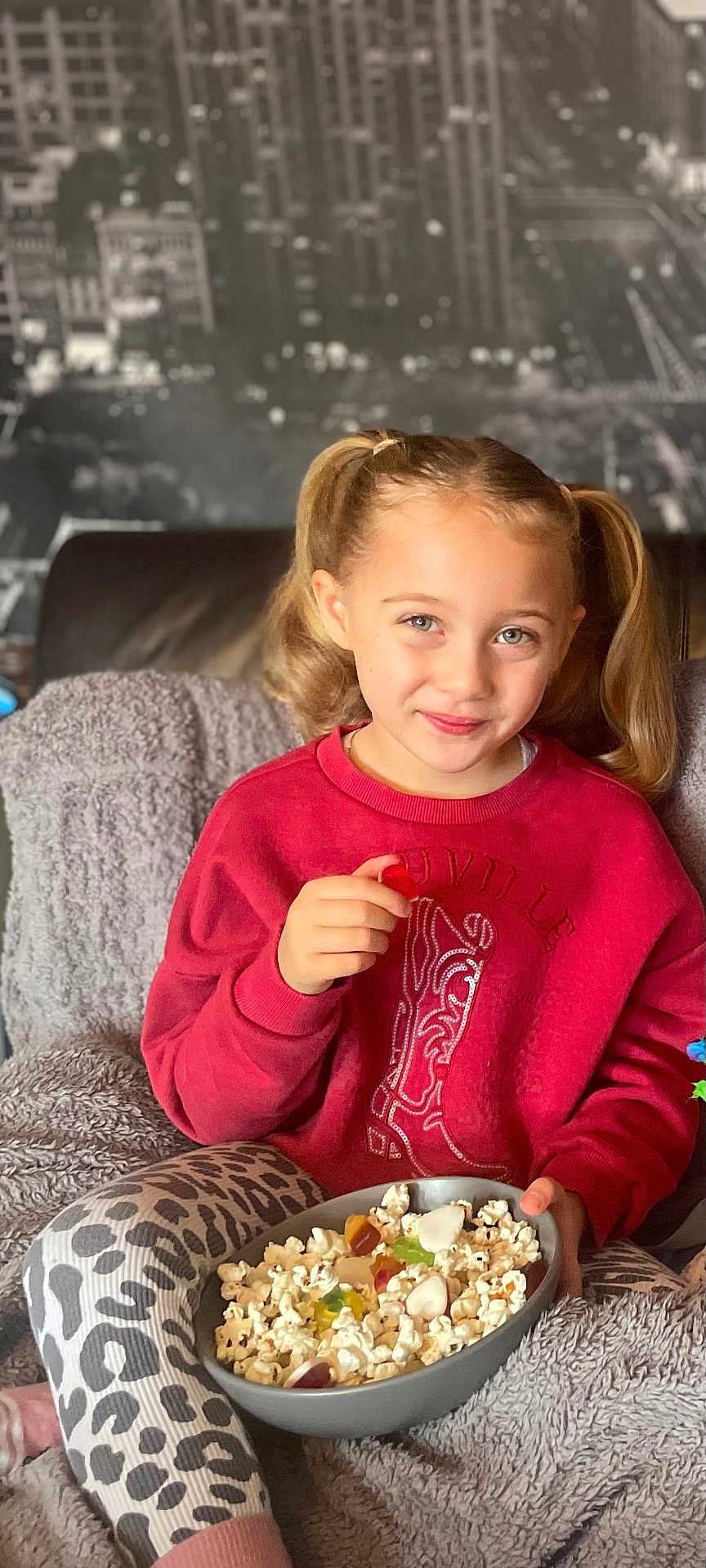 Zeliha joined the competition — help win amazing prizes! child, girl, pigtails, red_sweatshirt, leopard_print_leggings, popcorn, gummy_candies, bowl, blanket, couch, snacking, smiling, indoor, cozy, portrait, happy, casual_clothing, urban_background, seated, food