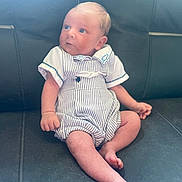 Bobby is registered to the contest to win money with this photo: baby, infant, child, sitting, romper, striped_clothing, leather_couch, indoor, cute, small, skin, head, face, legs, feet, hand, curious, looking_away, person, young