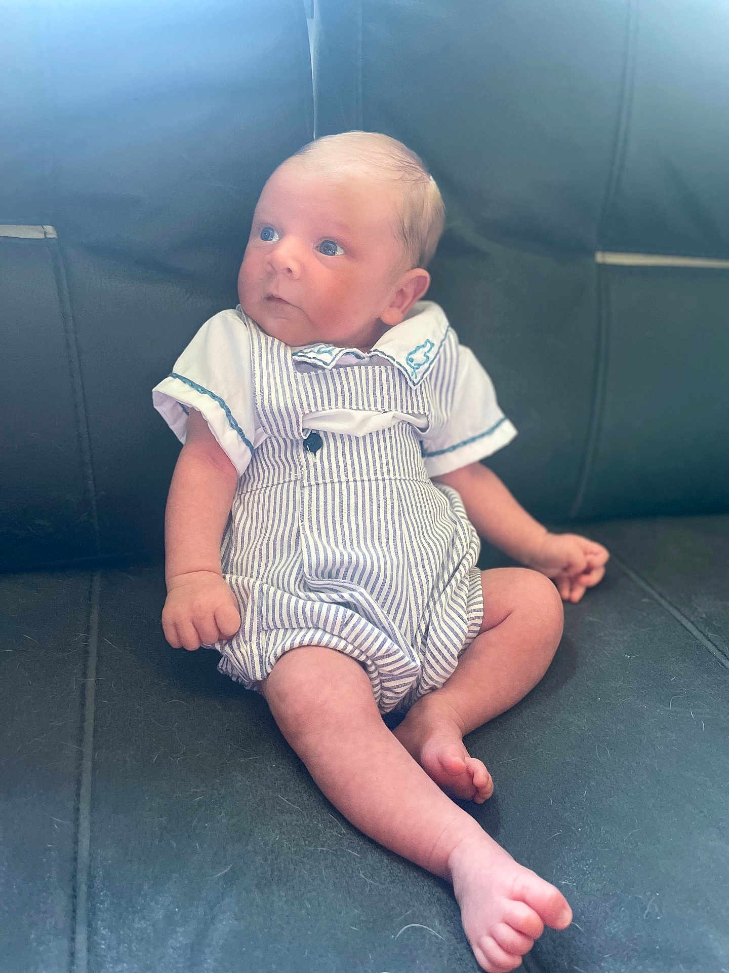 Bobby is registered to the contest to win money with this photo: baby, infant, child, sitting, romper, striped_clothing, leather_couch, indoor, cute, small, skin, head, face, legs, feet, hand, curious, looking_away, person, young
