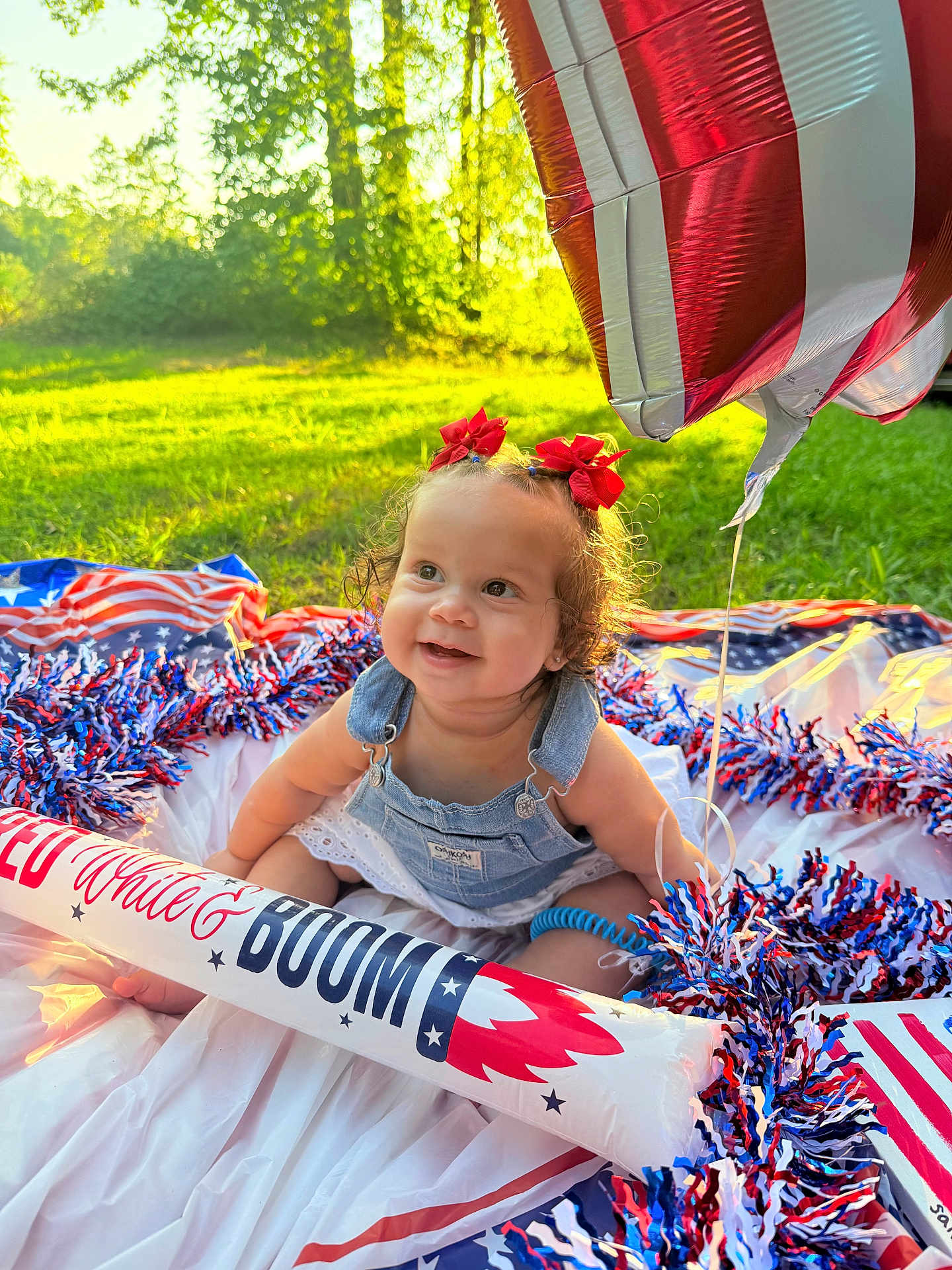 Sanova is registered to the contest to win money with this photo: baby, child, smiling, outdoor, grass, sunlight, balloon, red_bows, denim_overalls, patriotic, decorations, pom_poms, celebration, summer, happy, cute, nature, sitting, festive, portrait