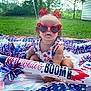 baby, child, sunglasses, heart_shaped, red_bows, patriotic, blanket, outdoor, grass, summer, celebration, red_white_blue, inflatable_rocket, festive, cute, smiling, holiday, fun, person, portrait