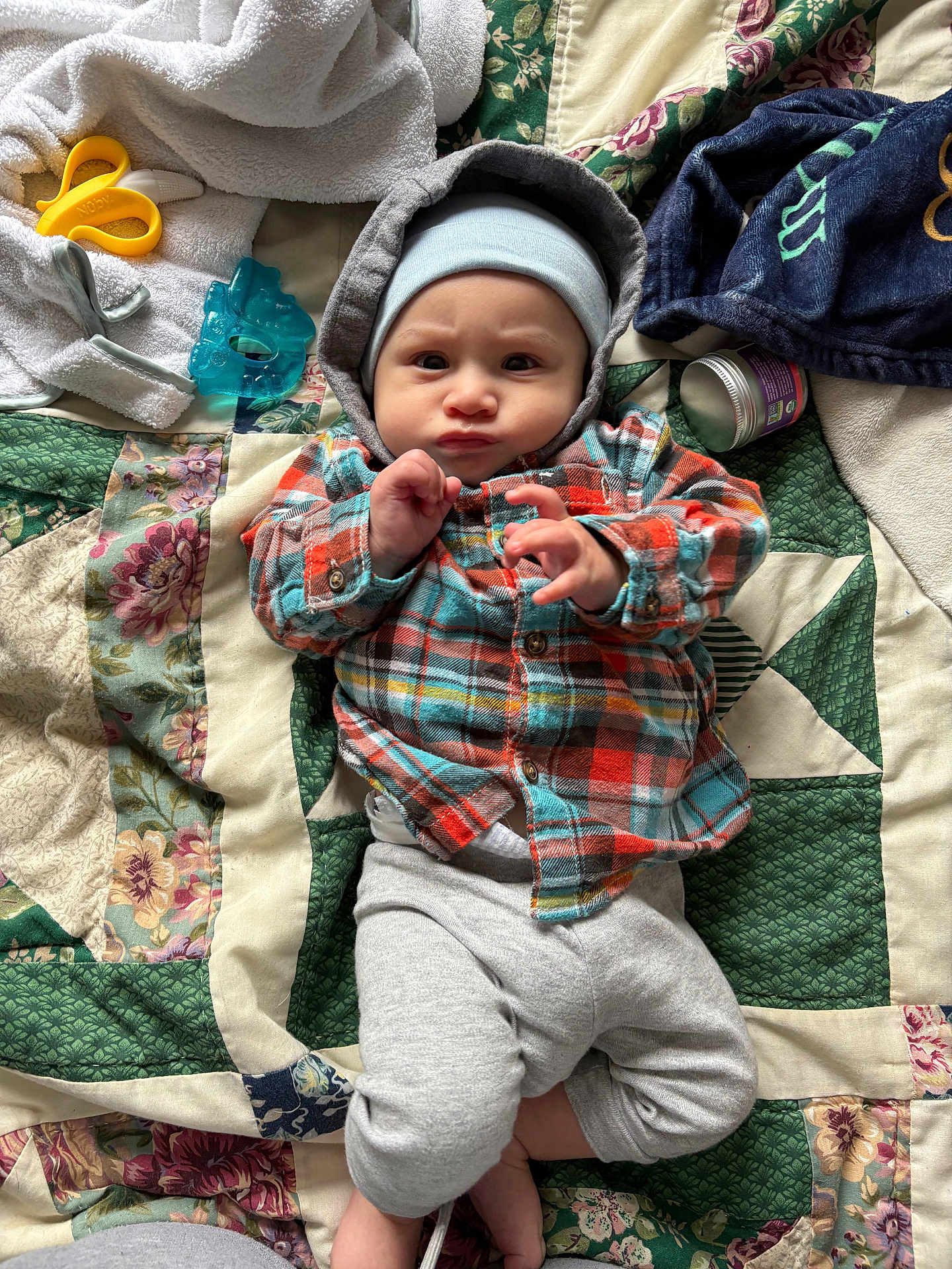 Alejandro is registered to the contest to win money with this photo: baby, child, plaid_shirt, hood, hat, quilt, blanket, toy, teether, container, indoor, laying_down, cute, curious_expression, infant, clothing, fabric, pattern, hands, feet