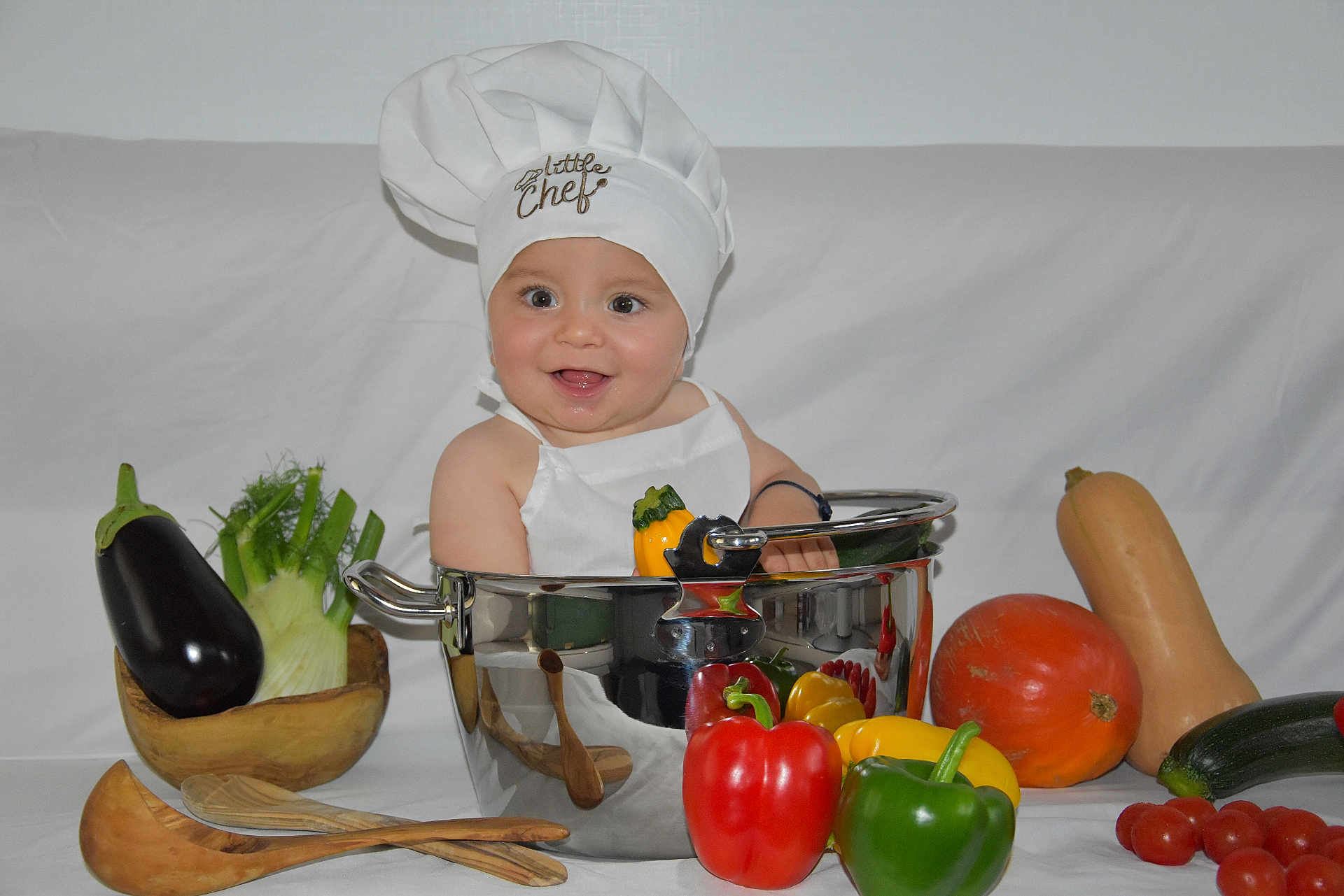 Dean participe au concours pour gagner de l'argent avec cette photo : apron, baby, bell_pepper, chef_hat, cherry_tomatoes, child, cooking_pot, eggplant, green_pepper, infant, portrait, pumpkin, red_pepper, smiling, stainless_pot, studio_background, vegetables, wooden_spoons, yellow_pepper, zucchini