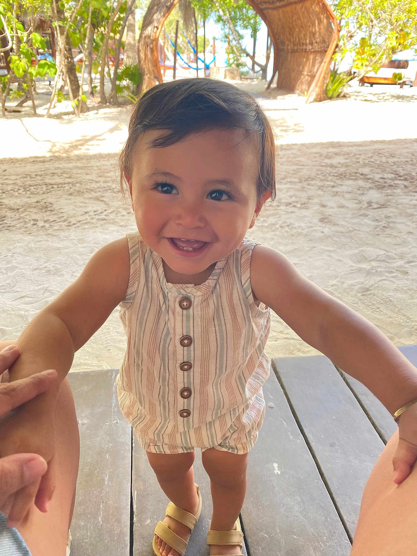 Gael is registered to the contest to win money with this photo: toddler, child, smiling, holding_hands, sand, beach, wooden_deck, sunlight, outdoor, trees, summer_clothing, stripes, sandals, happy, cute, portrait, nature, vacation, baby, playful