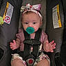 Madalyn is registered to the contest to win money with this photo: baby, child, car_seat, pacifier, headband, floral, safety_harness, infant, cute, pink_clothing, portrait, indoors, seat, hands, face, person, young_child, closeup, strapped_in, expression