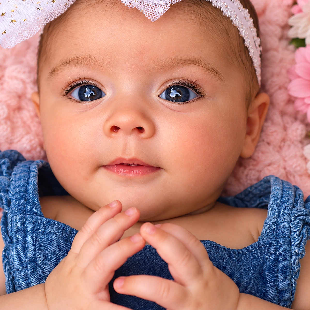 Evelynn is registered to the contest to win money with this photo: baby, infant, child, portrait, close_up, face, blue_eyes, wide_eyes, looking_at_camera, hands, fingers, headband, lace, blue_clothing, pink_flowers, floral_background, pink_blanket, lying_down, indoor, photography