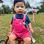 Sergio joined the competition — help win amazing prizes! toddler, child, outdoor, grass, tricycle, red_overalls, blue_shirt, face, person, vehicle, wheel, toy, nature, sky, cloud, tree, play, sitting, expression, summer