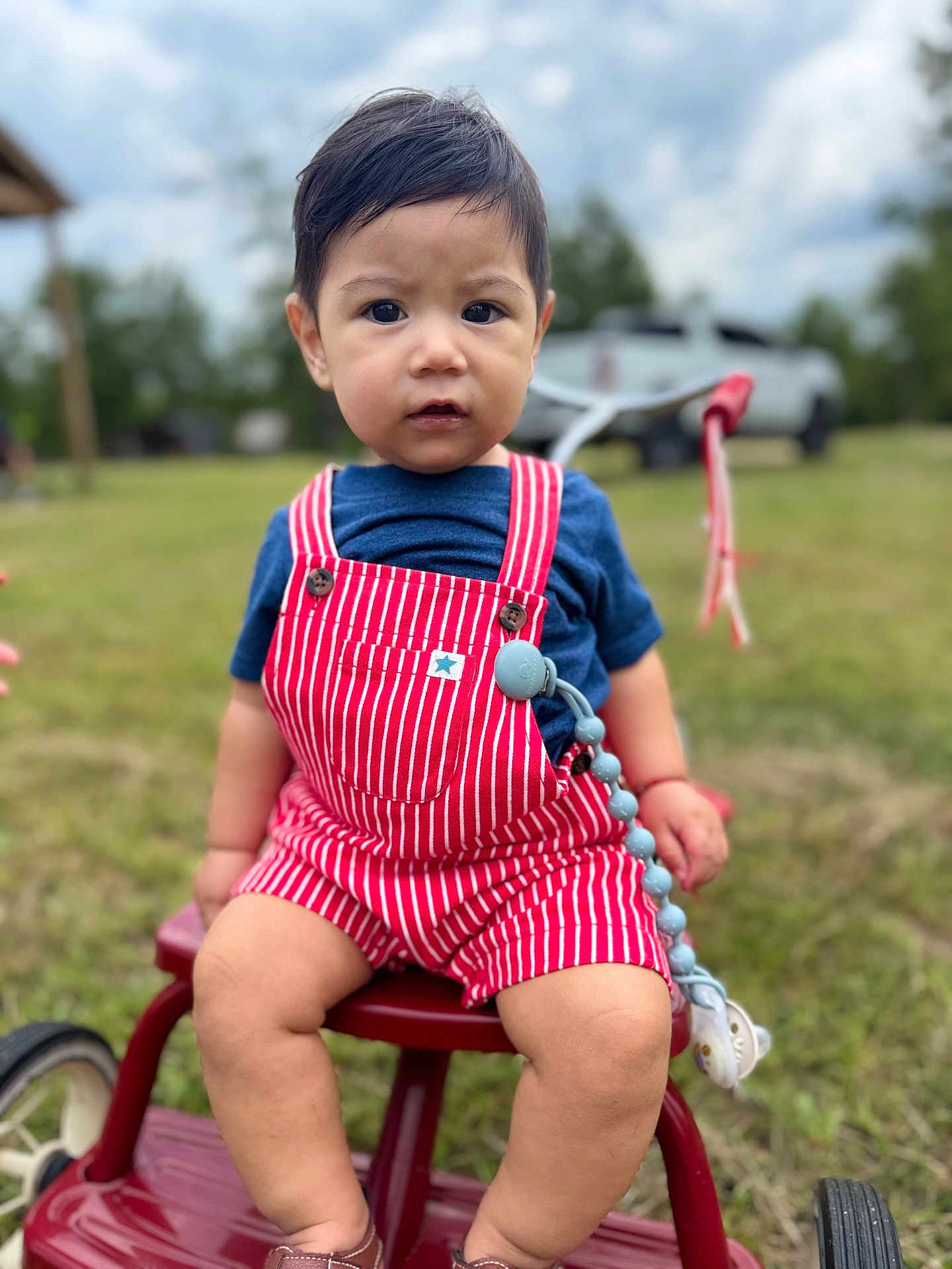 Sergio joined the competition — help win amazing prizes! toddler, child, outdoor, grass, tricycle, red_overalls, blue_shirt, face, person, vehicle, wheel, toy, nature, sky, cloud, tree, play, sitting, expression, summer