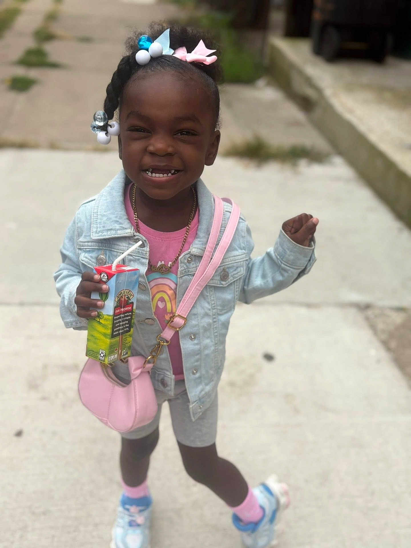 Nala is registered to the contest to win money with this photo: child, girl, smiling, juice_box, denim_jacket, pink_purse, hair_beads, bows, sidewalk, sneakers, shorts, pink_socks, casual_clothing, outdoor, happy, posing, person, accessories, cute, playful