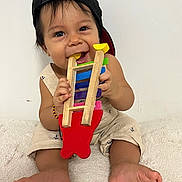 Nicolas joined the competition — help win amazing prizes! baby, child, toy, wooden_toy, cap, black_cap, smiling, cute, playing, sitting, barefoot, bracelet, colorful, indoor, white_background, happy, infant, chewing, clothing, person