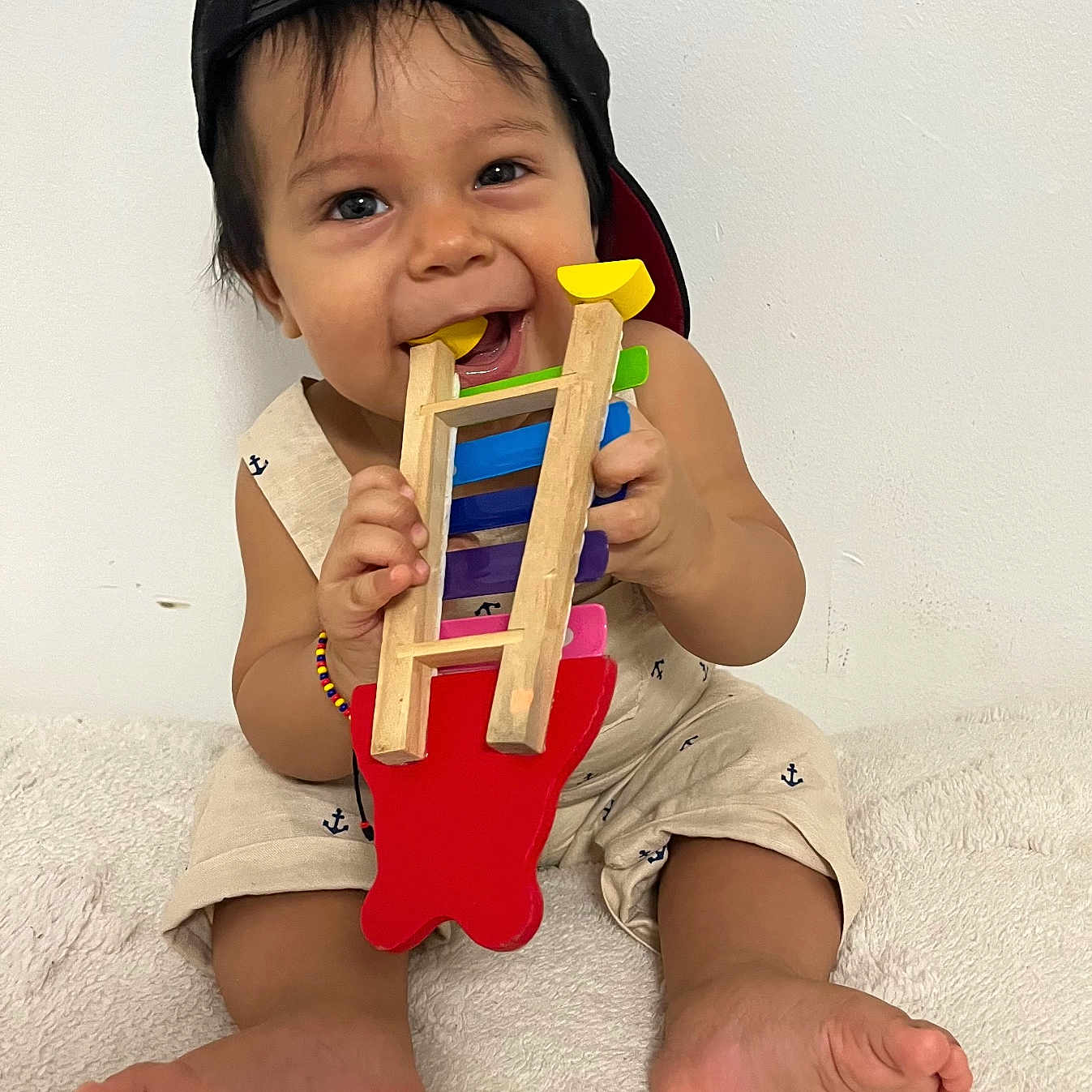 Nicolas joined the competition — help win amazing prizes! baby, barefoot, black_cap, bracelet, cap, chewing, child, clothing, colorful, cute, happy, indoor, infant, person, playing, sitting, smiling, toy, white_background, wooden_toy