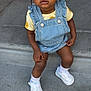 braided_hair, casual_clothing, child, concrete_steps, curious_expression, cute, daylight, denim_overalls, fashion, hair_clips, outdoor, portrait, sitting, small_earrings, sneakers, sunflower_pattern, toddler, urban, white_socks, young_child