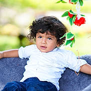 Rayansh is registered to the contest to win money with this photo: child, toddler, curly_hair, white_shirt, blue_pants, blanket, outdoor, greenery, flowers, red_flowers, relaxed, portrait, nature, cute, young_child, person, sitting, casual, daylight, expression
