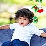 child, toddler, curly_hair, white_shirt, blue_pants, blanket, outdoor, greenery, flowers, red_flowers, relaxed, portrait, nature, cute, young_child, person, sitting, casual, daylight, expression