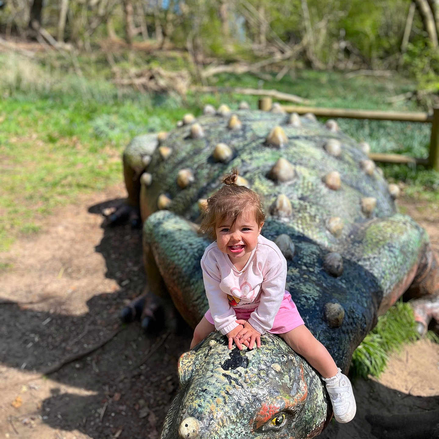 Daisy is registered to the contest to win money with this photo: adventure, casual_clothing, child, dinosaur_sculpture, grass, happy, long_sleeve_shirt, nature, outdoor, park, pink_skirt, playful, playground, smiling, statue, sunlight, toddler, toy, trees, white_shoes