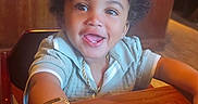 Legacy is registered to the contest to win money with this photo: arm, background, bracelet, child, clothing, curly_hair, face, furniture, happy, indoor, jewelry, lighting, person, portrait, restaurant, seat, smiling, table, toddler, wood