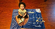 Legacy is registered to the contest to win money with this photo: baby, child, milestone_blanket, baseball, months, curly_hair, sneakers, pouting, flooring, indoor, cute, sitting, clothing, person, portrait, wood_floor, smile, number, toy, playful