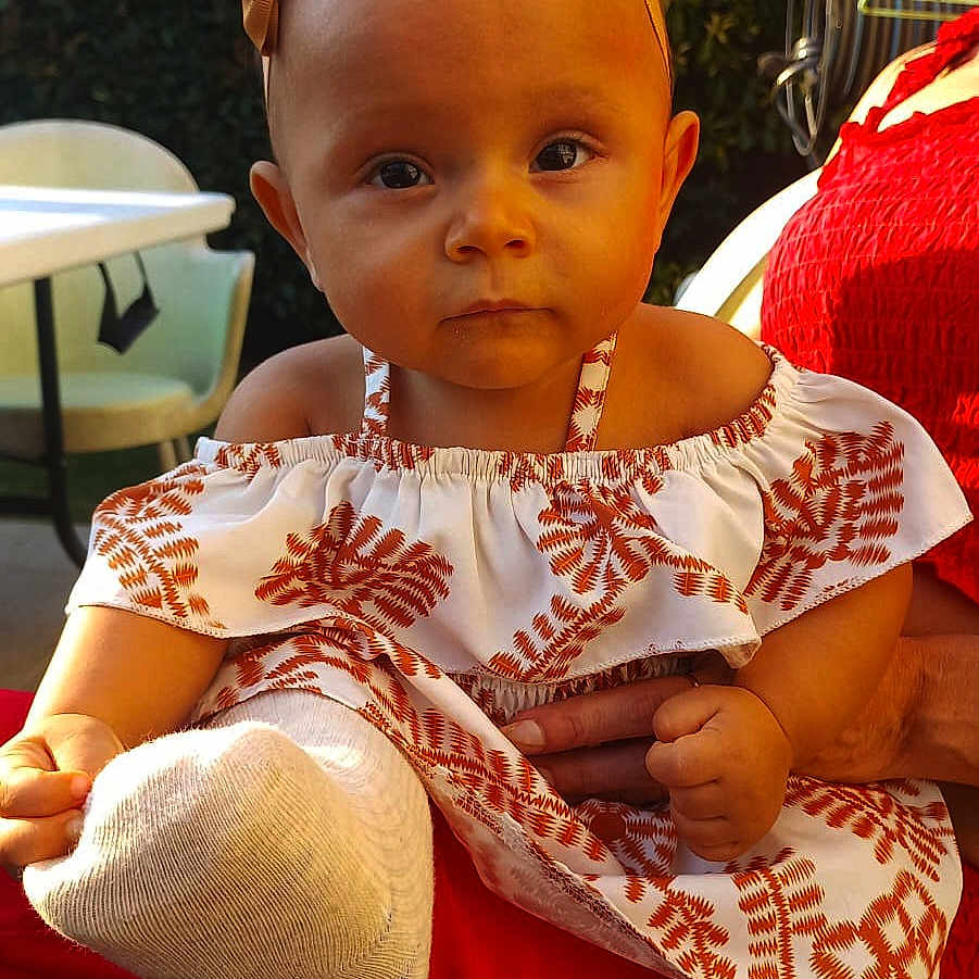 Mia participe au concours pour gagner de l'argent avec cette photo : baby, bow, chair, child, curious, dress, face, foot, greenery, hand, headband, outdoor, patterned_clothing, person, portrait, red_clothing, sitting, sock, sunlight, table
