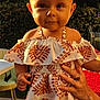 baby, child, dress, headband, outdoor, sunlight, garden, greenery, person, hands, patterned_clothing, smiling, cute, portrait, female, young_child, holding, warm_light, nature, background_blur
