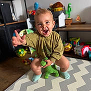 Morgan is registered to the contest to win money with this photo: toddler, child, toy, dinosaur, green_sweater, socks, zigzag_mat, indoor, playroom, smiling, happy, furniture, shelf, colorful_toys, wooden_floor, person, young_child, cute, playful, expression