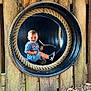 toddler, child, smile, blue_clothing, stick, black_tube, rope, wooden_fence, wood_chips, outdoor, playground, sneakers, happy, play, person, daylight, nature, fun, circular_frame, casual