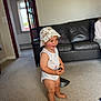 Morgan is registered to the contest to win money with this photo: toddler, child, white_onesie, bucket_hat, barefoot, carpet, living_room, black_leather_couch, smile, happy, indoor, person, baby, toy, floor, doorway, window, natural_light, home, cheek