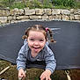 child, pigtails, tongue_out, trampoline, playful, springs, gray_sweatshirt, outdoor, grass, stone_wall, smiling, happy, young_child, person, fun, nature, expression, tongue, holding, casual_clothing